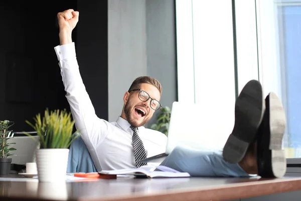 Knappe zakenman zittend met benen op tafel en houden arm opgevoed en uitdrukken vreugde in kantoor — Stockfoto