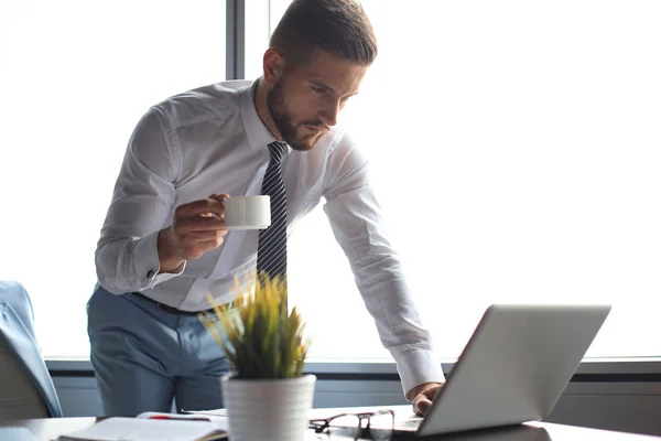 Вдумчивый молодой бизнесмен пользуется компьютером и пьет кофе во время работы в офисе — стоковое фото