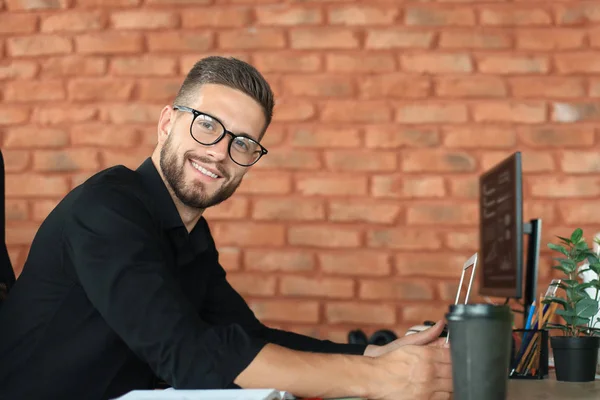 Joven hombre de negocios está utilizando el ordenador portátil mientras está sentado en la oficina, mirando a la cámara, sonriendo — Foto de Stock