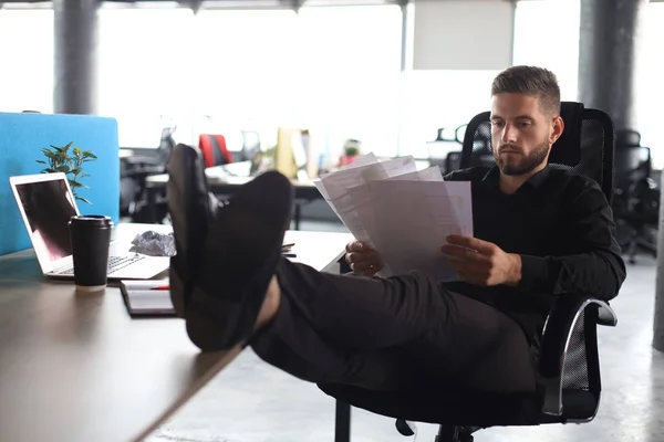 Udany przedsiębiorca bada dokumenty z uważnym i skoncentrowanym wyglądem, nogami na biurku — Zdjęcie stockowe