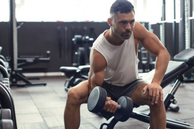 Formda ve kaslı bir adam spor salonunda dambıllarla kol egzersizi yapıyor..