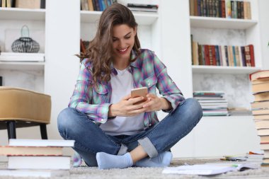 Genç kız öğrenci eğitim e dönmeden önce sosyal medyayı kontrol ediyor, rahat iç mekana karşı yerde oturuyor, kitap yığınıyla çevrili.