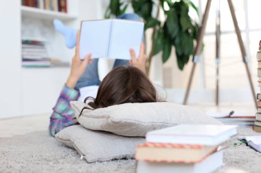 Evde okuyan, üniversite sınavlarına hazırlanan, rahat iç mekana karşı yerde yatan, kitap yığınıyla çevrili genç kız öğrenci.