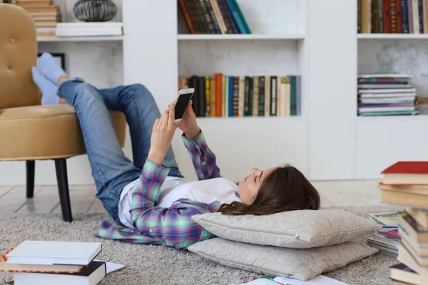 Junge Studentin checkt soziale Medien, bevor sie wieder studiert, liegt auf dem Boden vor dem gemütlichen häuslichen Interieur, umgeben von einem Stapel Bücher. — Stockfoto