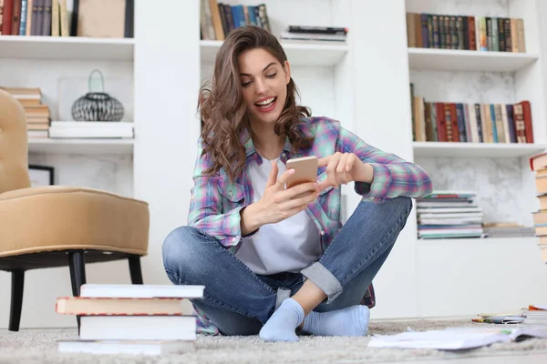 Joven estudiante revisando las redes sociales antes de volver a estudiar, sentada en el suelo contra el acogedor interior doméstico, rodeada de un montón de libros . —  Fotos de Stock