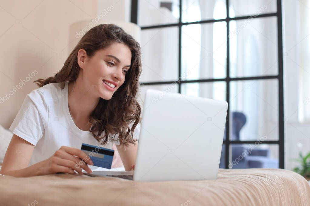 Happy natural brunette using credit card and laptop in bedroom.