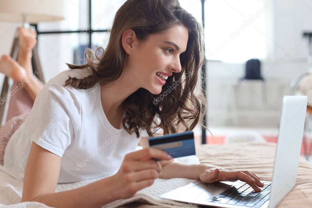 Happy natural brunette using credit card and laptop in bedroom.