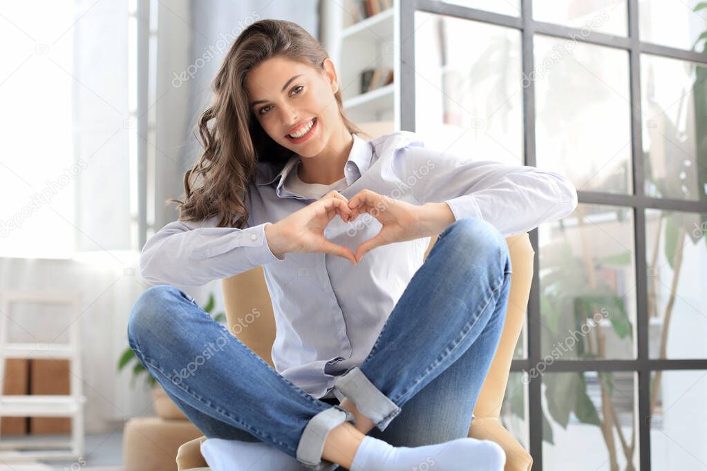 Smiling young woman is showing heart gesture with two hands and looking at camera sitting on armchair.