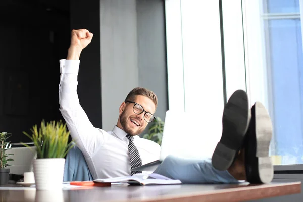 Hombre de negocios guapo sentado con las piernas en la mesa y manteniendo el brazo levantado y expresando alegría en la oficina — Foto de Stock