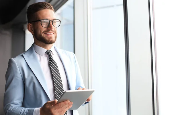 Сучасний бізнесмен у формальному одязі з використанням цифрового планшета, стоячи біля вікна в офісі — стокове фото