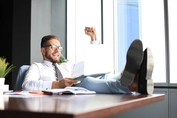 Przystojny biznesmen siedzi z nogami na stole i trzymając ramię wychował i wyrażając radosny w biurze — Zdjęcie stockowe