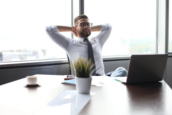 Jovem homem de negócios moderno mantendo as mãos atrás da cabeça e sorrindo enquanto sentado no escritório — Fotografia de Stock