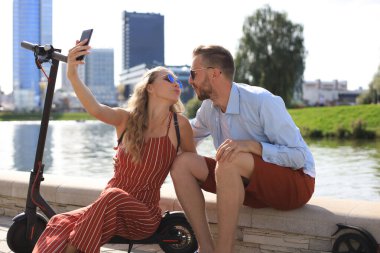 Güzel bir çift, elektrikli scooter kullanırken eğleniyor, araba kullanmaya ara veriyor, nehir kıyısında oturuyor, selfie çekiyor.