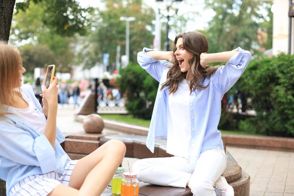 Счастливые девушки друзья фотографируют друг друга на скамейке в городе . — стоковое фото