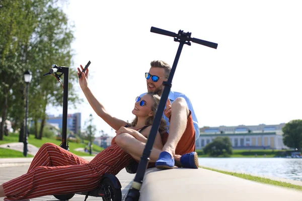 Schönes Paar, das Spaß am Fahren von Elektrorollern hat, eine Pause vom Autofahren macht, am Ufer sitzt, Selfie macht — Stockfoto