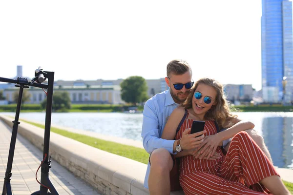 Krásný pár baví řízení elektrický skútr, dává si pauzu od řízení, sedí na břehu řeky, pomocí mobilního telefonu — Stock fotografie