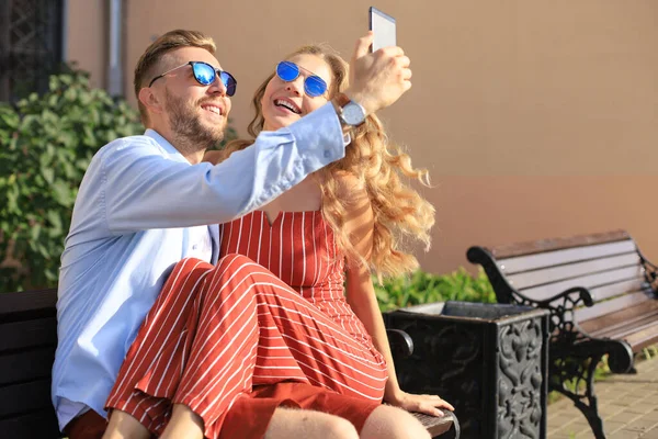 Romantický mladý pár v letním oblečení s úsměvem a přičemž selfie zatímco sedí na lavičce ve městě ulici — Stock fotografie