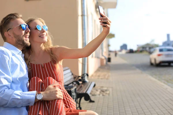 Romântico jovem casal em roupas de verão sorrindo e tomando selfie enquanto sentado no banco na rua da cidade — Fotografia de Stock