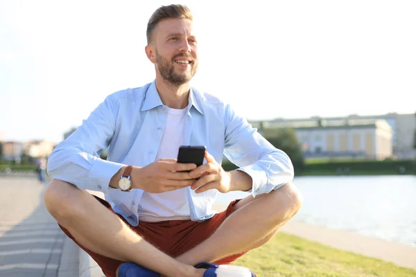 Boldog jóképű fiatalember ült a padon a szabadban, és használ smartphone — Stock Fotó