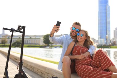 Güzel bir çift, elektrikli scooter kullanırken eğleniyor, araba kullanmaya ara veriyor, nehir kıyısında oturuyor, selfie çekiyor.