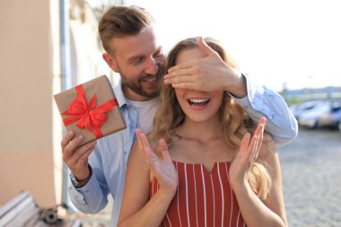 Mutlu genç bir çiftin portresi hediye kutusuyla yan yana dururken sarılıyor, erkek kadının gözlerini kapatıyor.