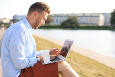 Yakışıklı adam şehirde vakit geçiriyor, bankta oturup kahve içerken dizüstü bilgisayar kullanıyor.