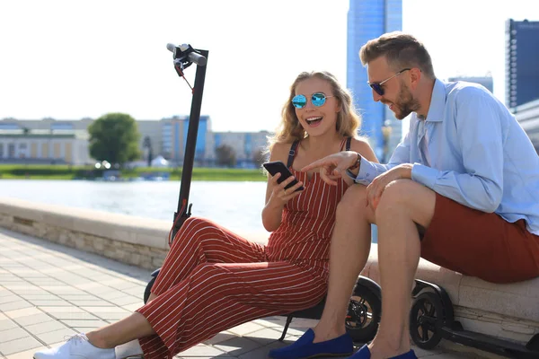 Charmant couple s'amusant à conduire un scooter électrique, prenant une pause de la conduite, assis sur la rive, en utilisant un téléphone mobile — Photo