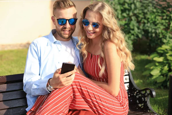 Romántica pareja joven en ropa de verano sonriendo y usando el móvil mientras se sienta en el banco en la calle de la ciudad — Foto de Stock
