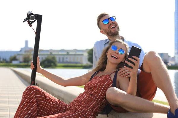 Preciosa pareja divirtiéndose conduciendo scooter eléctrico, tomando un descanso de conducir, sentado en la orilla del río, tomando selfie — Foto de Stock