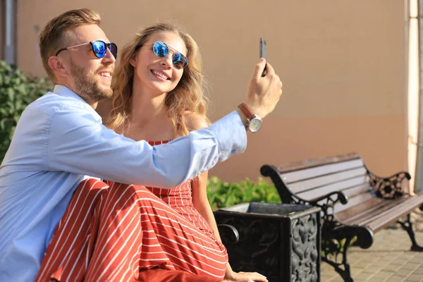 Romântico jovem casal em roupas de verão sorrindo e tomando selfie enquanto sentado no banco na rua da cidade — Fotografia de Stock