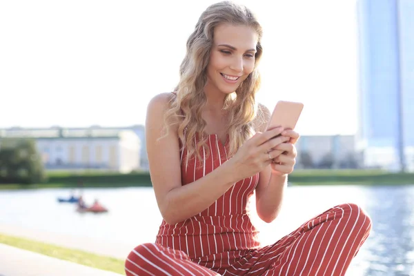 Portré csinos fiatal nő ül a folyóparton lábak keresztbe a nyári nap folyamán, okostelefon használatával — Stock Fotó