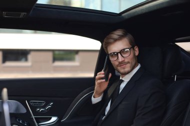 Takım elbiseli yakışıklı, akıllı telefonla konuşan ve arabada otururken dizüstü bilgisayar kullanarak çalışan yakışıklı kendine güvenen bir işadamı.