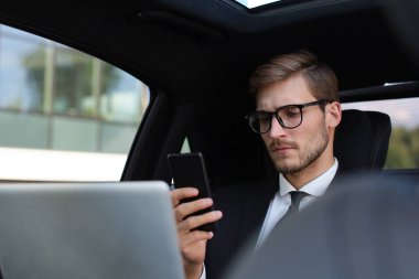 Arabada otururken ve dizüstü bilgisayar kullanırken onun akıllı telefon bakarak tam takım elbise yakışıklı kendinden emin adam.