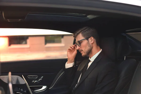 Lüks arabada otururken gözlük elini tutmak düşünceli kendinden emin işadamı. — Stok fotoğraf