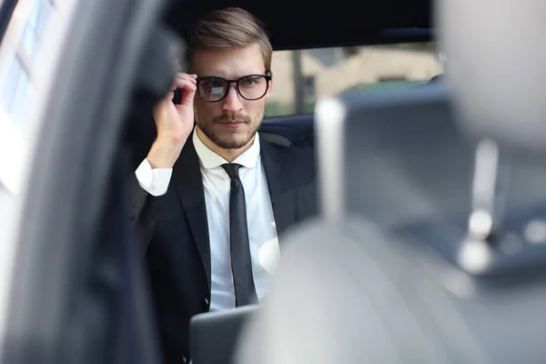 Thoughtful confident businessman keeping hand on glasses while sitting in the luxe car. — Stock Photo, Image