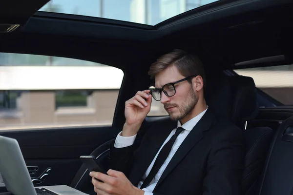 Bonito homem confiante em terno completo olhando para seu telefone inteligente enquanto sentado no carro e usando laptop . — Fotografia de Stock