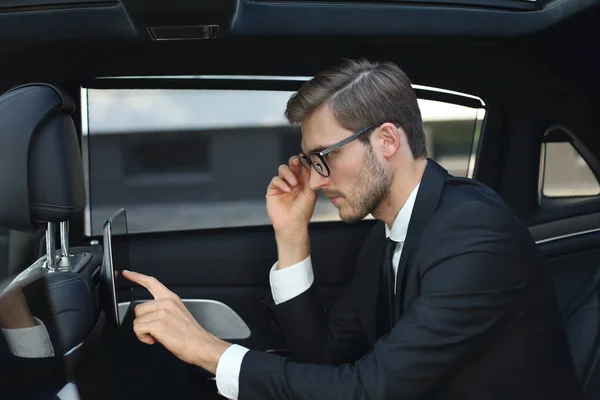 Pensativo joven empresario sentado en el coche de lujo y el uso de su tableta . — Foto de Stock