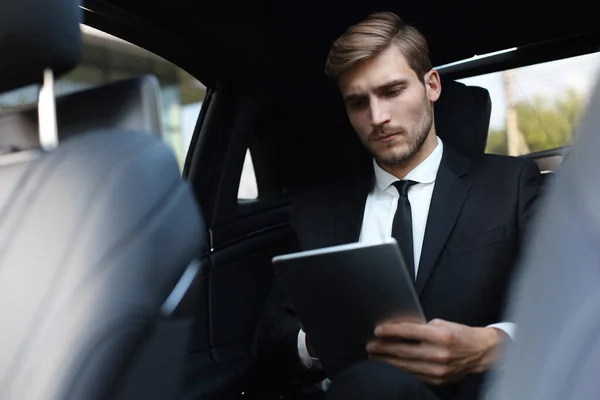 Giovane uomo d'affari premuroso seduto in macchina di lusso e utilizzando il suo tablet . — Foto Stock