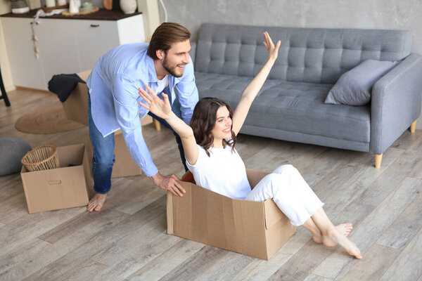 Happy couple is having fun with cardboard boxes in new house at moving day, woman riding sitting in cardboard box while man pushing it.