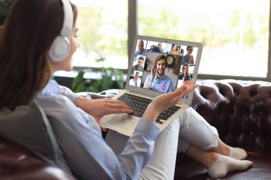 Kanepeye uzanmış kulaklıklı bir iş kadını, kişisel tecrit ve karantina sırasında iş arkadaşlarıyla online brifing hakkında görüntülü konuşma yapıyor. Ofisteki iş arkadaşlarıyla webcam grup konferansı.