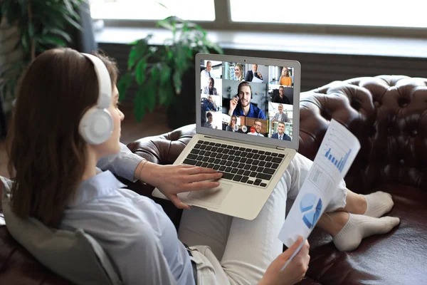 Geschäftsfrau mit Kopfhörer auf Sofa liegend spricht in Videokonferenz mit ihren Kollegen. Geschäftsteam arbeitet von zu Hause aus mit Laptop und diskutiert Finanzbericht seines Unternehmens. — Stockfoto