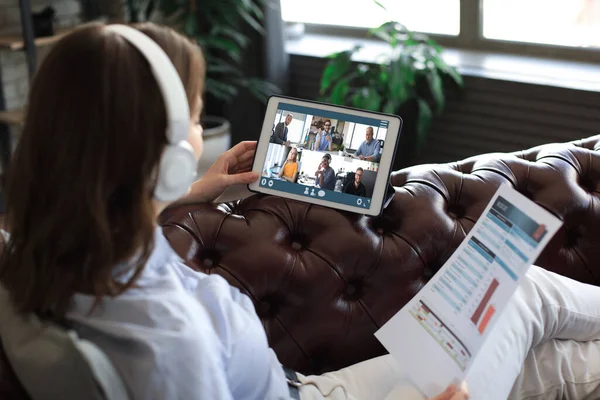 Geschäftsfrau mit Kopfhörer auf Sofa liegend spricht in Videokonferenz mit ihren Kollegen. Geschäftsteam arbeitet von zu Hause aus mit digitalem Tablet und diskutiert Finanzbericht seines Unternehmens. — Stockfoto