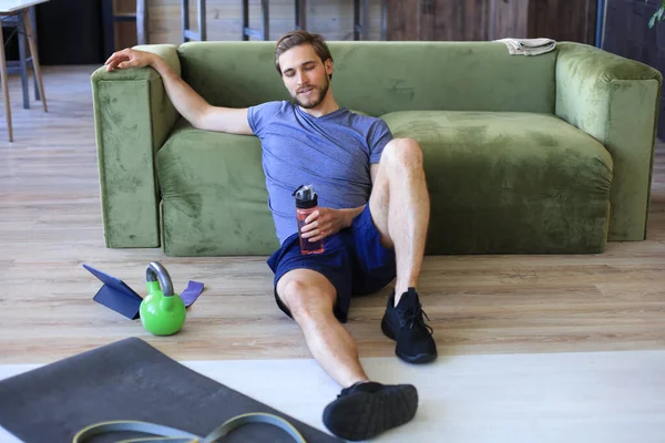 スポーツマンの休憩 休憩飲料水の生活室で床に座って運動をした後 自己分離中の家での罰金 — ストック写真