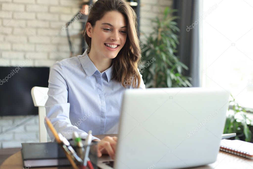 Smiling pretty woman sitting at table, looking at laptop screen. Happy entrepreneur reading message email with good news, chatting with clients online