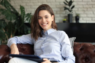 Oturma odasındaki kanepede oturan ve dijital tablet kullanan gülümseyen genç bayan.