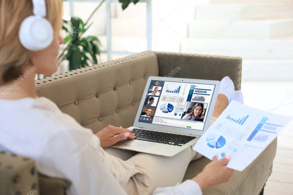 Business woman in headphones lying on sofa speak talking to her colleagues in video conference. Business team working from home using laptop, discussing financial report of their company.
