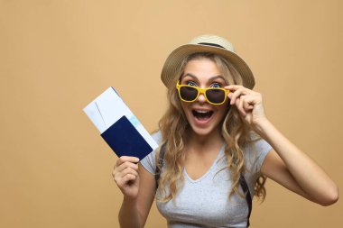 Güneş gözlüklü, pasaportlu, bej arka planda biletleri olan güzel sarışın kadın.