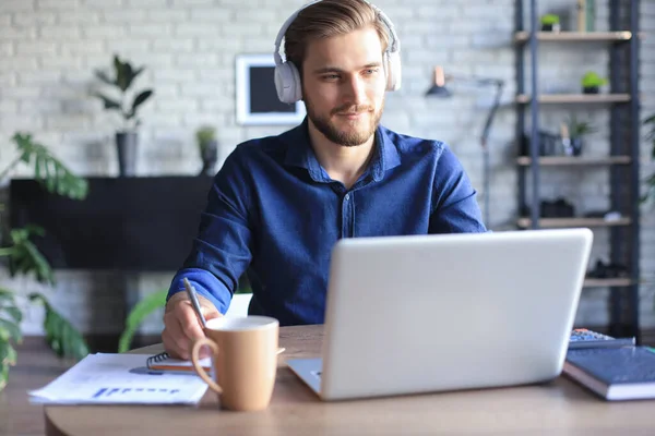 Confident businessman in earphones is writing notes or financial report while sitting at desk with laptop at home