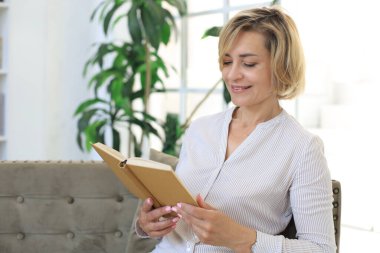 Çekici orta yaşlı kadın oturma odasındaki kanepede oturmuş kitap okurken gülümsüyor.