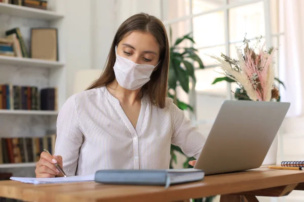 在自我隔离和隔离期间 戴着医疗防护面具的年轻女商人在家里的笔记本电脑旁工作 — 图库照片